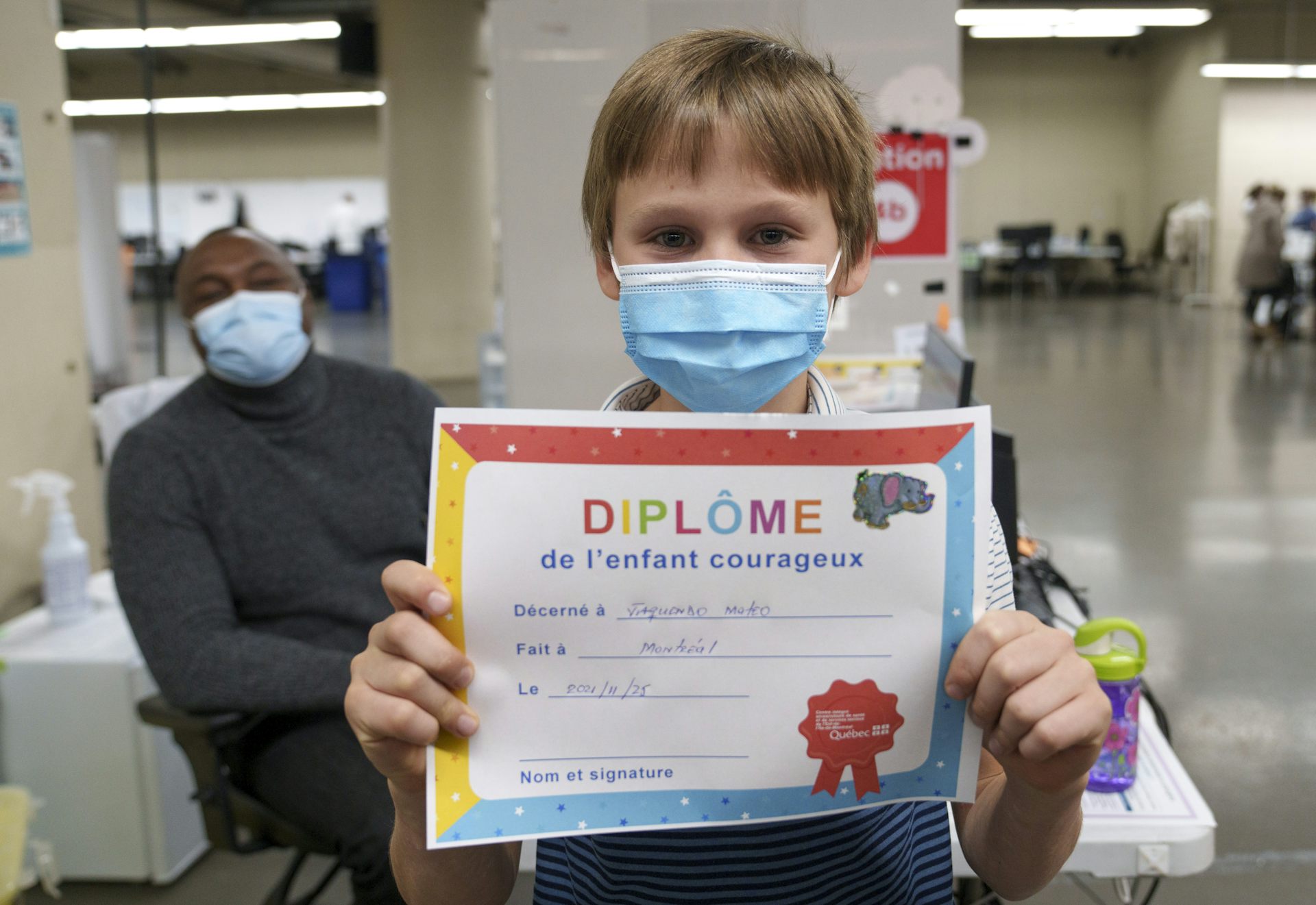 A little boy in a face mask holds up his French-language brave child diploma after getting vaccinated