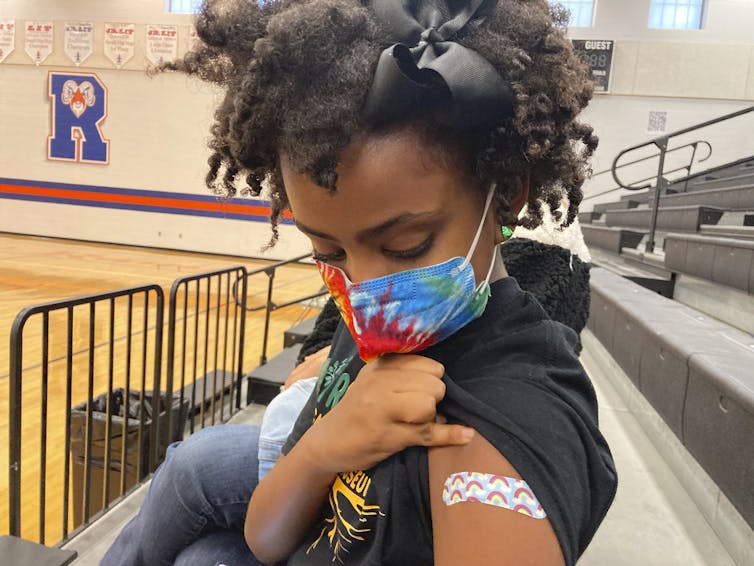 A Black girl wearing a face mask and a black hair ribbon holds her sleeve up to show the bandage over her vaccination site