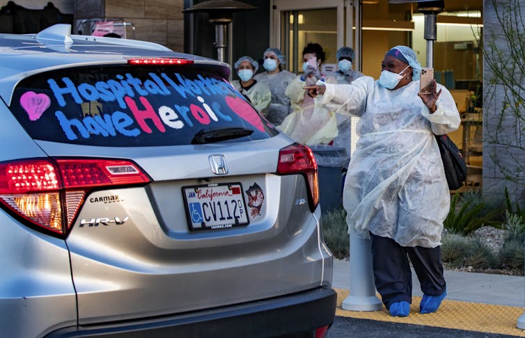 Women in surgical scrubs gesture at a car bearing the message hospital workers have heroic hearts