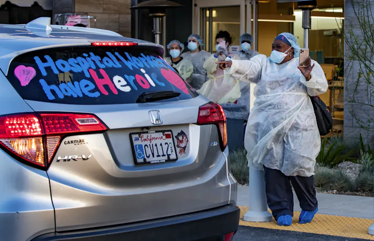 Women in surgical scrubs gesture at a car bearing the message hospital workers have heroic hearts