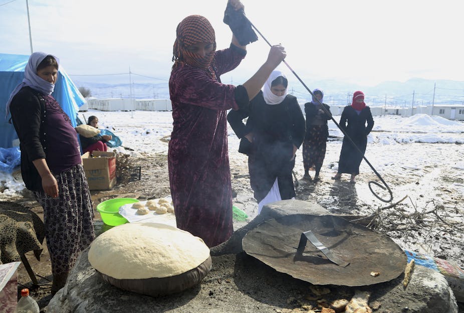 A woman in a headscarf joined by others bakes bread in front of a tent in an open field.