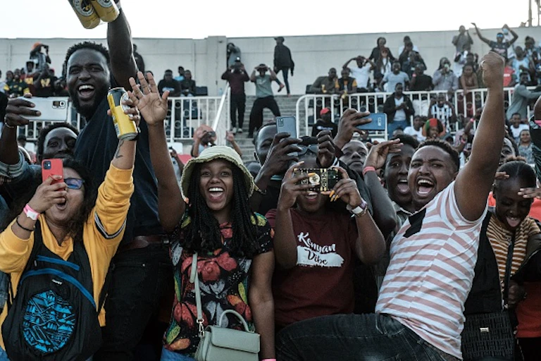Crowd of people with arms raised, smiling and holdings phones up, at a stadium
