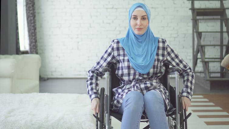 An interior shot of a woman wearing a blue headscarf and a checkered dress in a wheelchair.