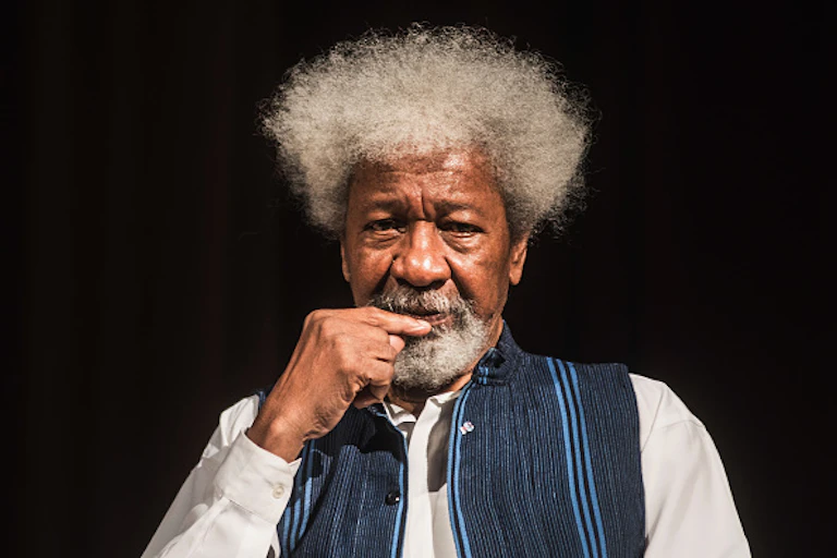 An elderly man with a shock of white hair holds a finger to his chin.