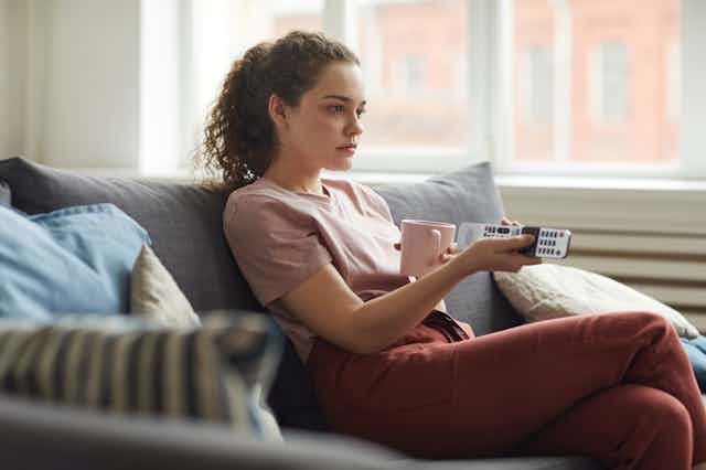 A young woman sits on the couch, holding a TV remote.