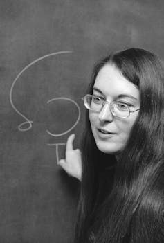A woman with glasses points to a chalkboard.