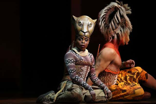 Two actors on stage, dressed in elaborate animal costumes - a woman looks to camera, sitting, a lion mask above her head.