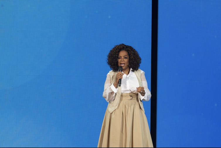 Oprah Winfrey, speaking into a hand-held microphone against a blue backdrop.