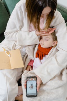 A woman cradling a toddler, with a phone in one hand consulting with a doctor, with her other hand on the child's forehead