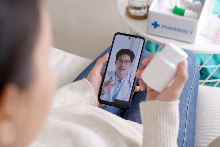 A woman's hands, one holding her phone speaking with a doctor, the other holding a bottle of medicine