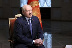 A man in a dark suit and a moustache sits in a red upholstered chair.