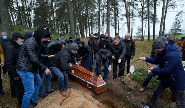 A group of people in dark jackets and jeans lower a casket into a grave in a forested area..