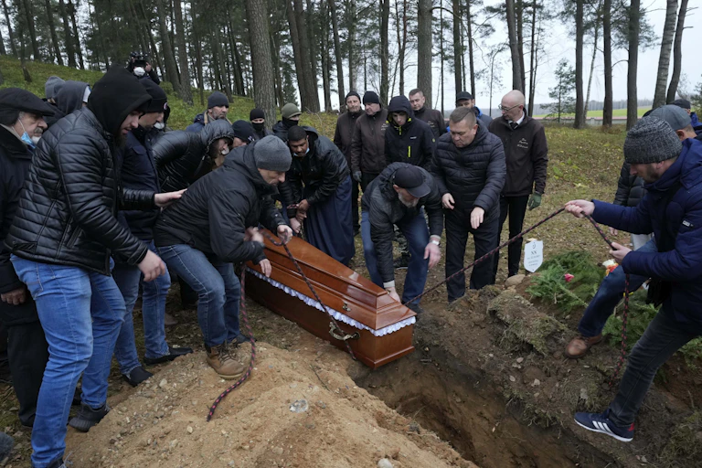 A group of people in dark jackets and jeans lower a casket into a grave in a forested area..