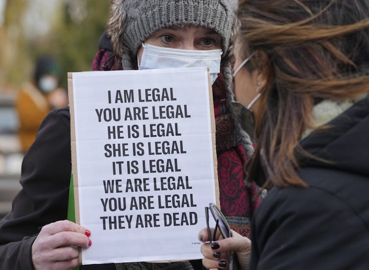 A woman in a wool hat and mask carries a sign saying 'I am legal, you are legal, he is legal, she is legal, it is legal, we are legal, you are legal, they are dead.'