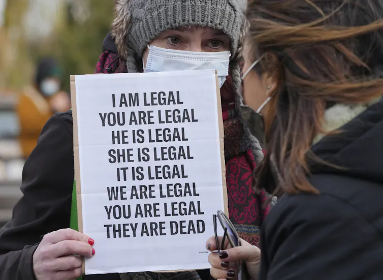 A woman in a wool hat and mask carries a sign saying 'I am legal, you are legal, he is legal, she is legal, it is legal, we are legal, you are legal, they are dead.'