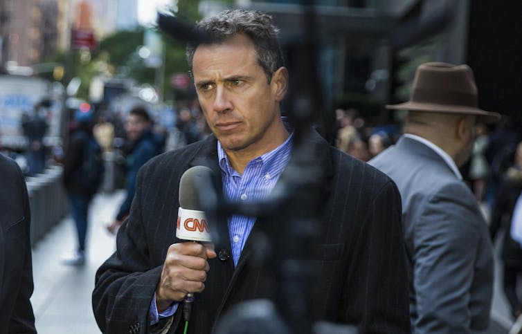 Chris Cuomo on a city sidewalk, talking into a microphone for a news report.