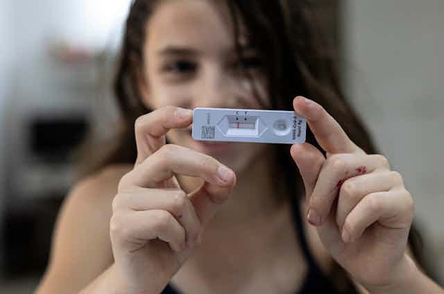 A girl out of focus in the background holding up a negative COVID-19 rapid antigen test in the foreground