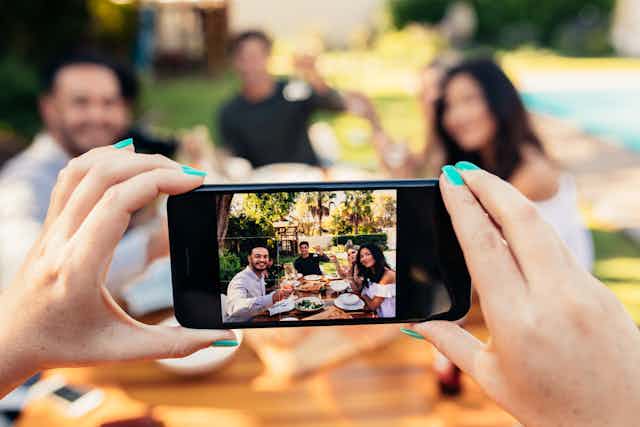Hands hold a smartphone taking a photo of a group of friends dining outdoors.