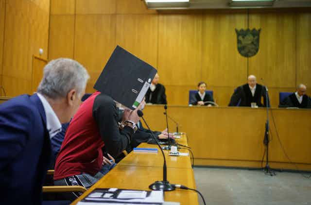 Scene in a German courtroom. The defendant, Taha al-Jumailly, hides his face behind a folder. He is flanked by lawyers. In the background are the judges.