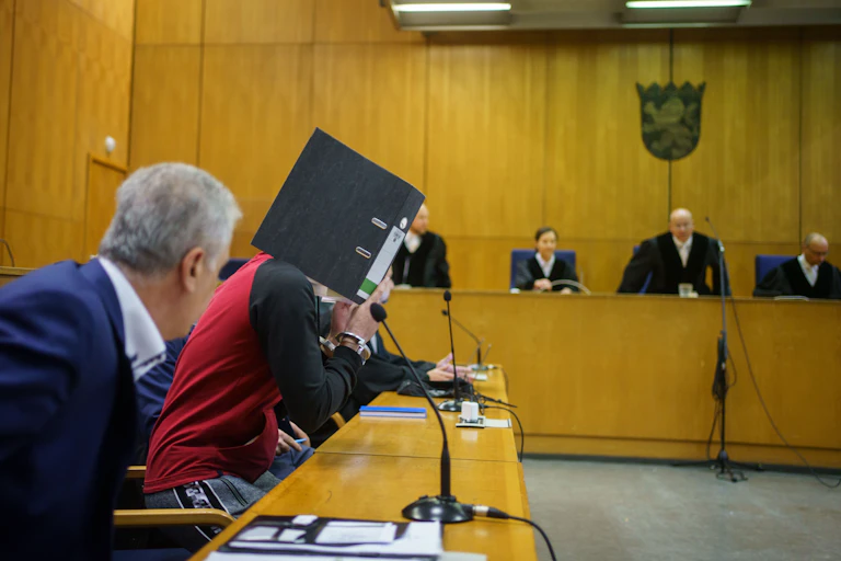Scene in a German courtroom. The defendant, Taha al-Jumailly, hides his face behind a folder. He is flanked by lawyers. In the background are the judges.