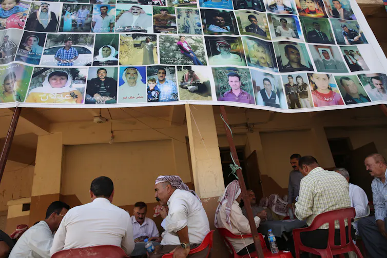 Yazidi men sit in a tea house in Dohuk, Northern Iraq. ABove them is a banner with the photographs of some of the Yazidi people kidnapped or sold into slavery since 2014.