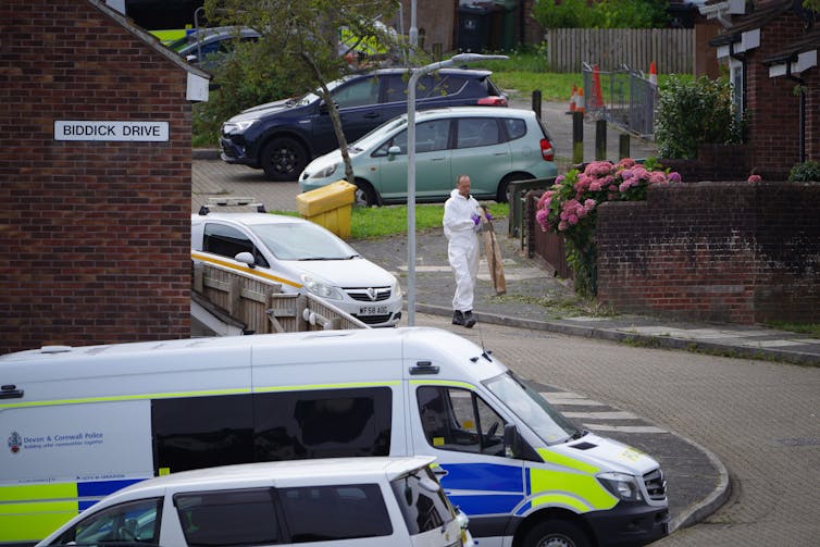 Man in white overalls walks down residential street with police van.