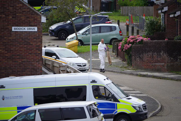 Man in white overalls walks down residential street with police van.