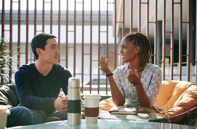 A woman and man sit at a table with coffees in front of them they are engaged in conversation.