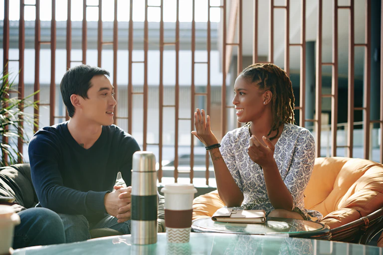 A woman and man sit at a table with coffees in front of them they are engaged in conversation.