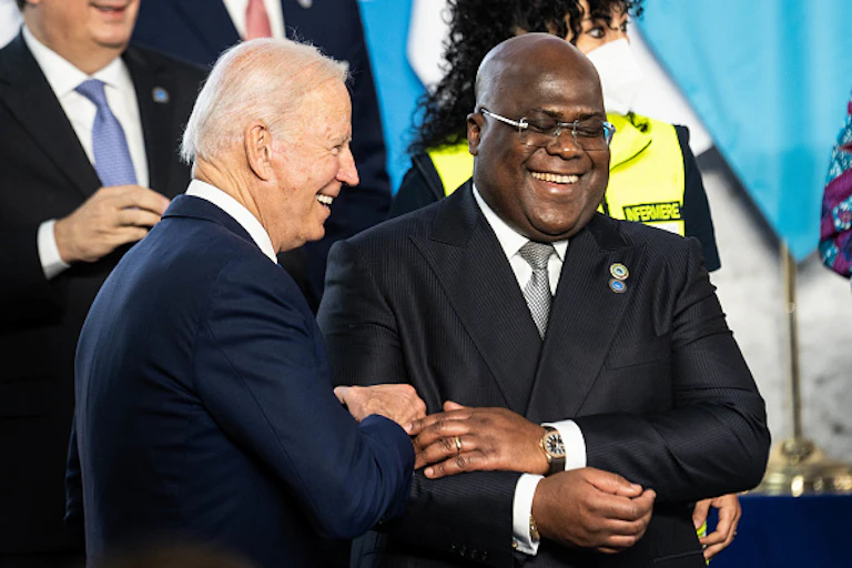 An elderly man in a suit touches the arm of another man in a suit as both of them smile
