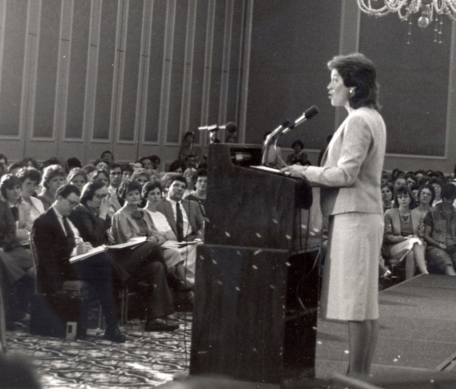 Woman with short hair, standing at a lectern, speaking to an audience..