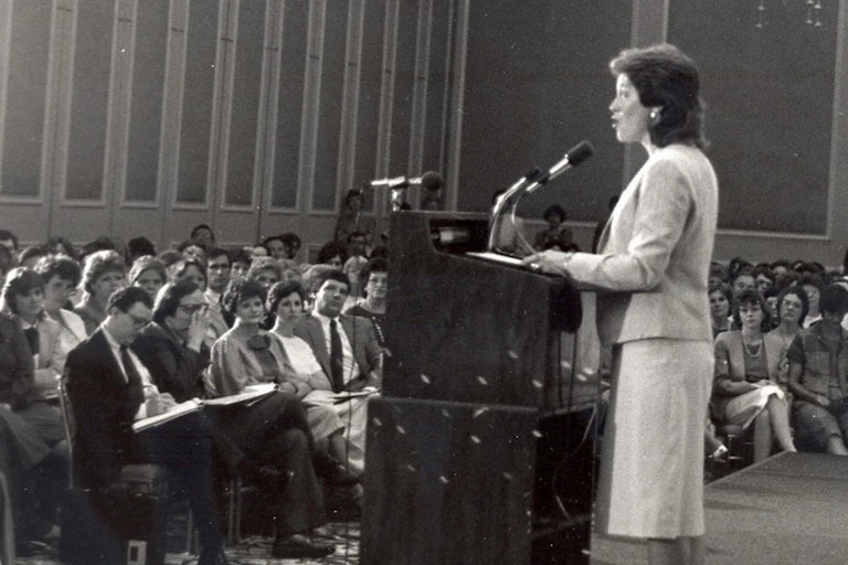 Woman with short hair, standing at a lectern, speaking to an audience..
