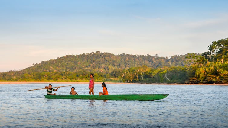 Four teenagers paddle a long green canoe in a tropical river.