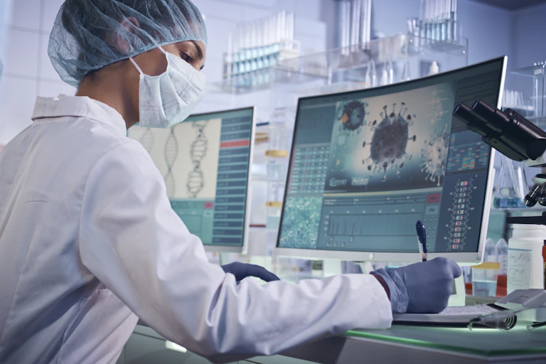 masked woman in white coat seated at computer with virus on screen