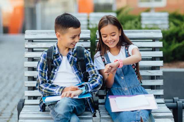 Two children sit on a bench looking at their smartwatches.