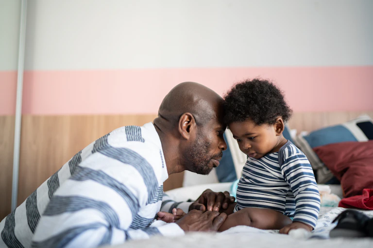 Father leans over to touch foreheads with baby sitting on a bed