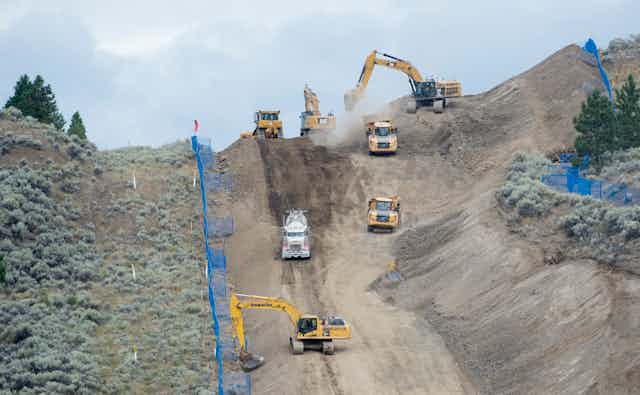 Backhoes and dump trucks excavate a hillside marked off by blue fences.