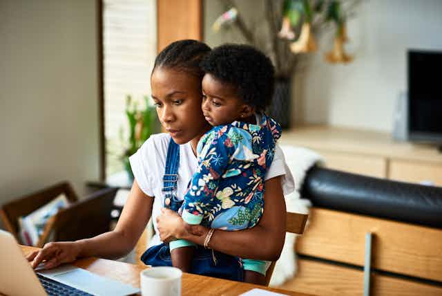 A Black mother works on her laptop while holding her child.
