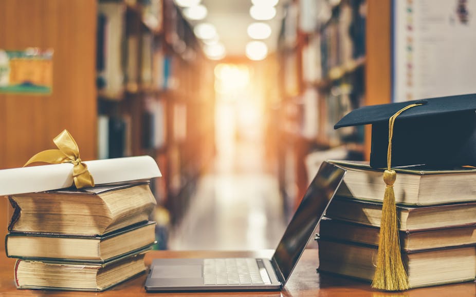 A laptop with two stacks of books on either side. On one stack there's an academic cap and on the other is an academic certificate. Blurred library background.