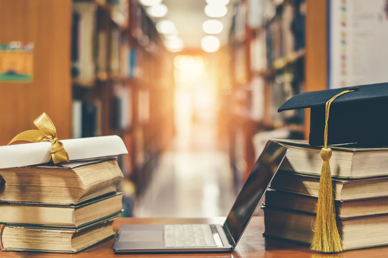 A laptop with two stacks of books on either side. On one stack there's an academic cap and on the other is an academic certificate. Blurred library background.