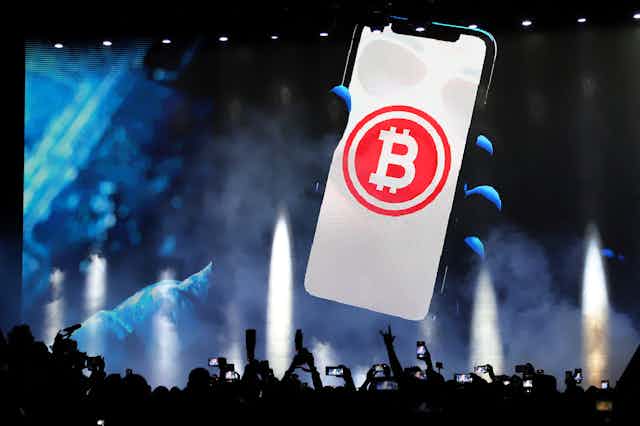 A bitcoin symbol on a giant screen with people in front of it waving their arms and taking photos.