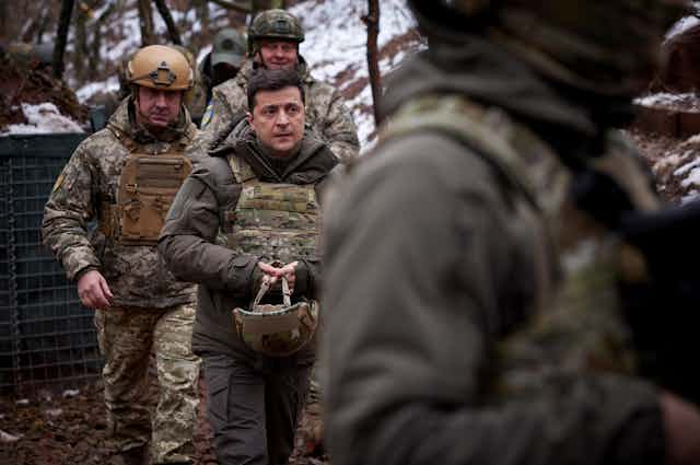 Ukrainian president, Volodymyr Zelensky, dressed in military camouflage and carrying a helmet. He is accompanied by military personnel and political aides.
