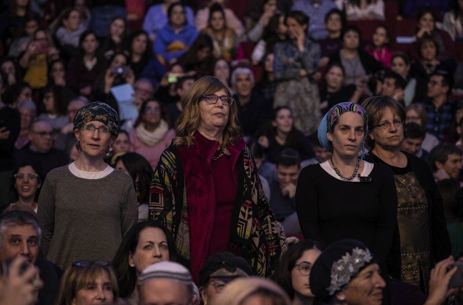 Three women, two of whom are wearing headscarves, stand in an auditorium full of women.