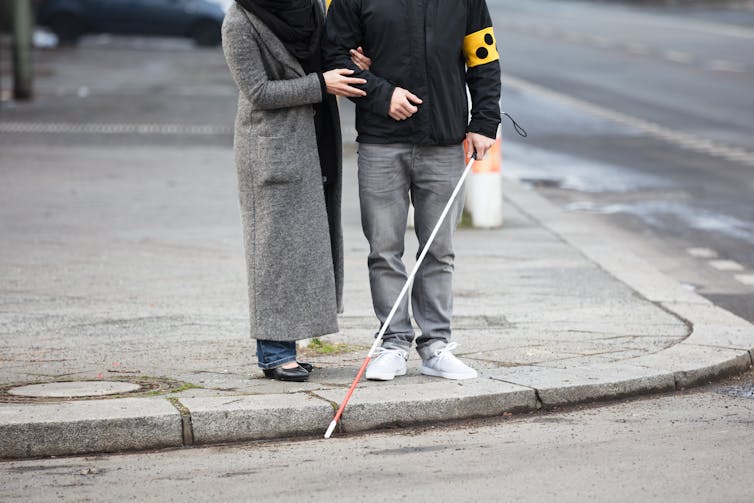 Woman holding the arm of a man, who is holding a white cane. Both are standing on a curb.