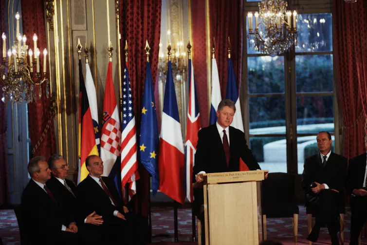 Former President Bill Clinton stands behind a lectern surrounded by leaders of several countries.