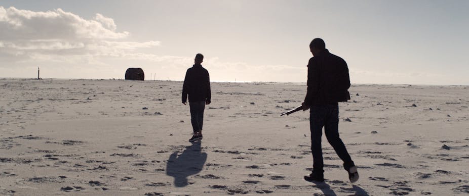 Two men walking on the beach, one holding a gun at the other's back