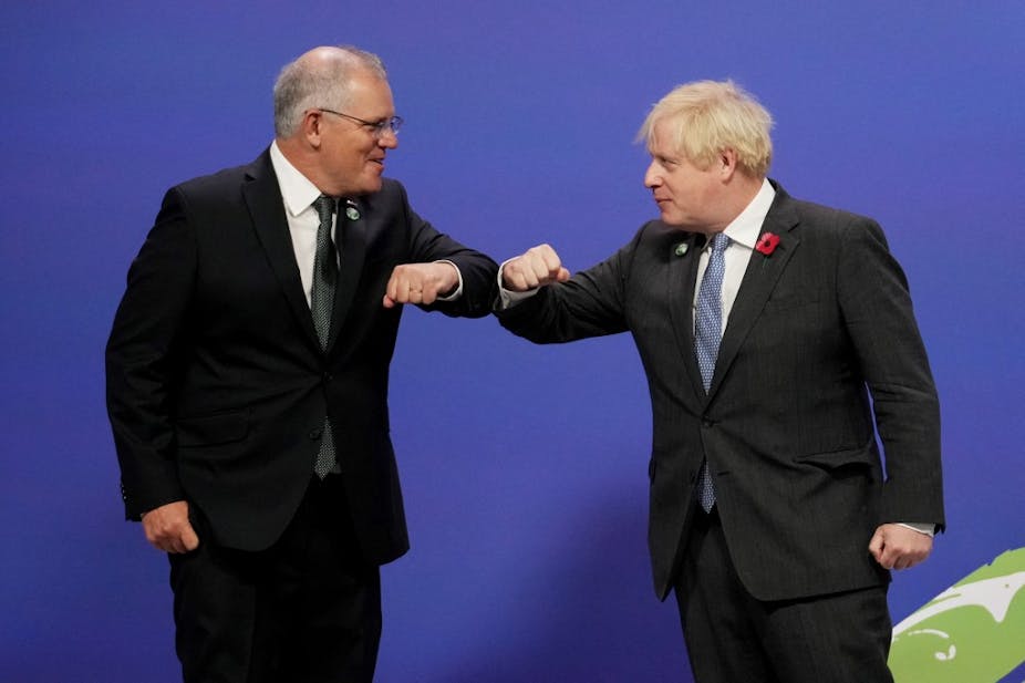 Boris Johnson et Scott Morrison, en costumes sombres sur fond bleu, se saluent du coude.