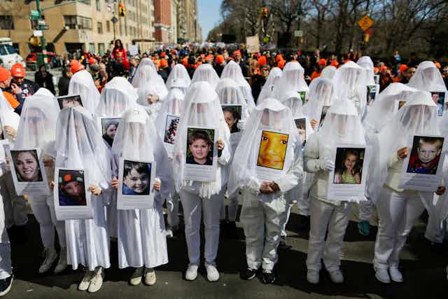 A row of people draped in white stand together holding close-up photos of people.