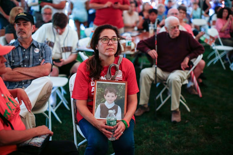 A woman in a red shirt sits among a group of people and holds a photo of a young boy.