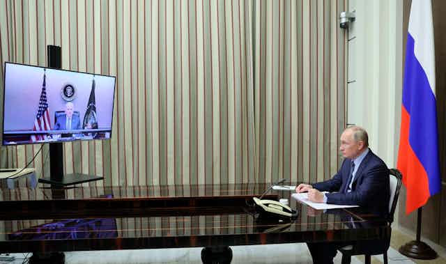 Putin sits at a table with a Russian flag behind him as he looks at a tv screen that displays biden sitting at a desk